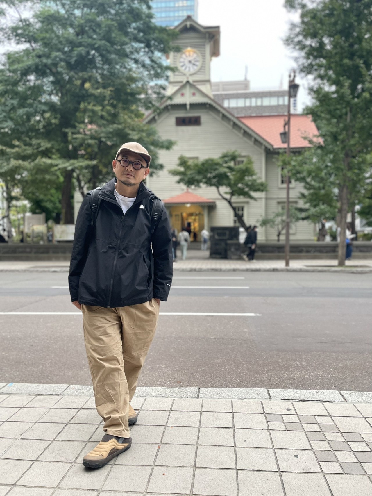 Shen Guang Tang director Jeff Liu, full-length portrait before the Sapporo Clock Tower: beige cap and round glasses, black jacket and khaki trousers, with the red-roofed wooden clock tower and street trees behind