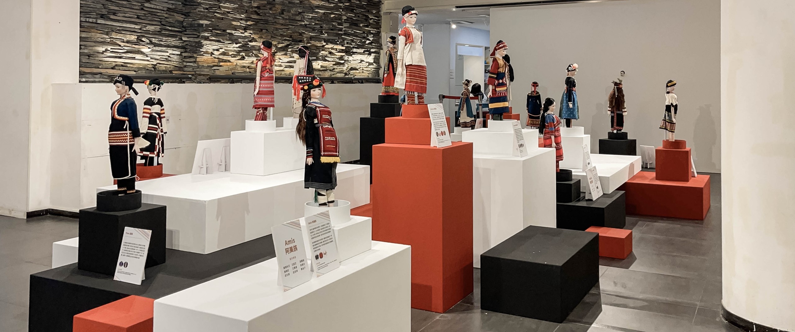 原住民族服飾展區:各族傳統服飾娃娃立於紅、白、黑三色高低錯落的塊體展台上,後方石牆與「阿美族 Amis」圖文說明卡,展台節奏明快而有秩序