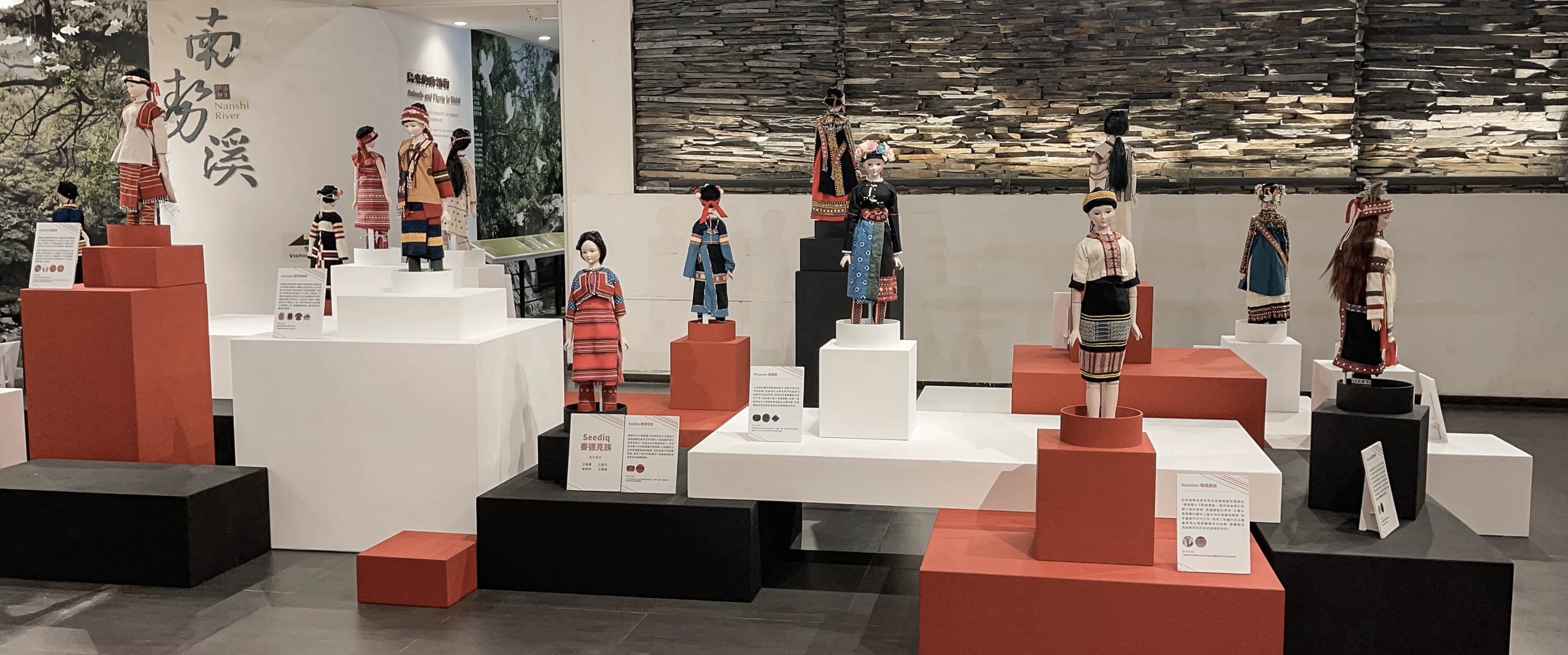 原住民族服飾展區全景:以「南勢溪 Nanshi River」為背景牆,賽德克、卑南、噶瑪蘭等各族服飾娃娃分立於紅白黑塊體展台,構成一條族群文化的動線