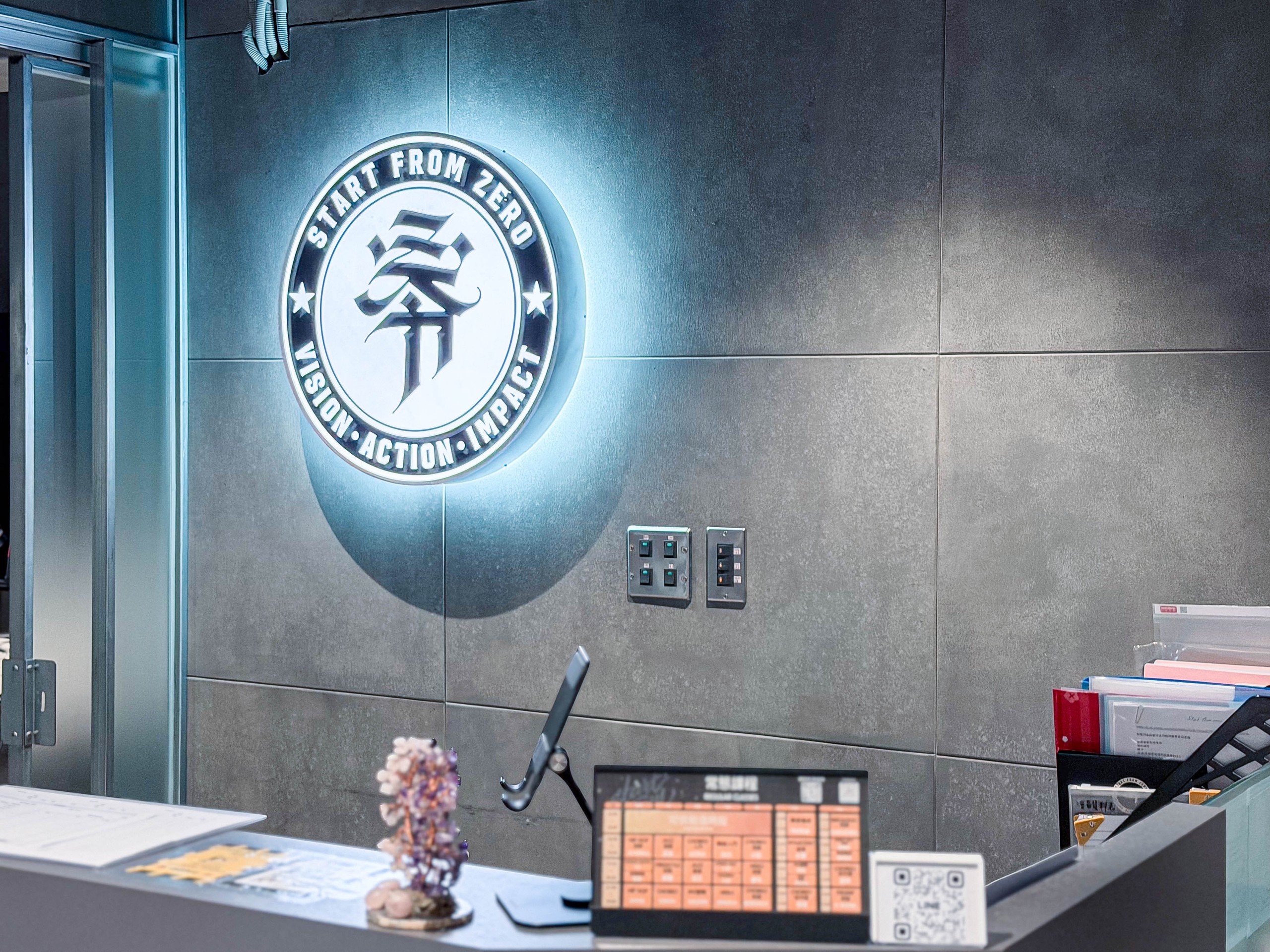 舞蹈教室櫃台牆:背光發亮的圓形「朵」字徽章貼附清水模牆面,下方金屬控制開關,旁邊玻璃隔間,工業冷調卻有儀式感