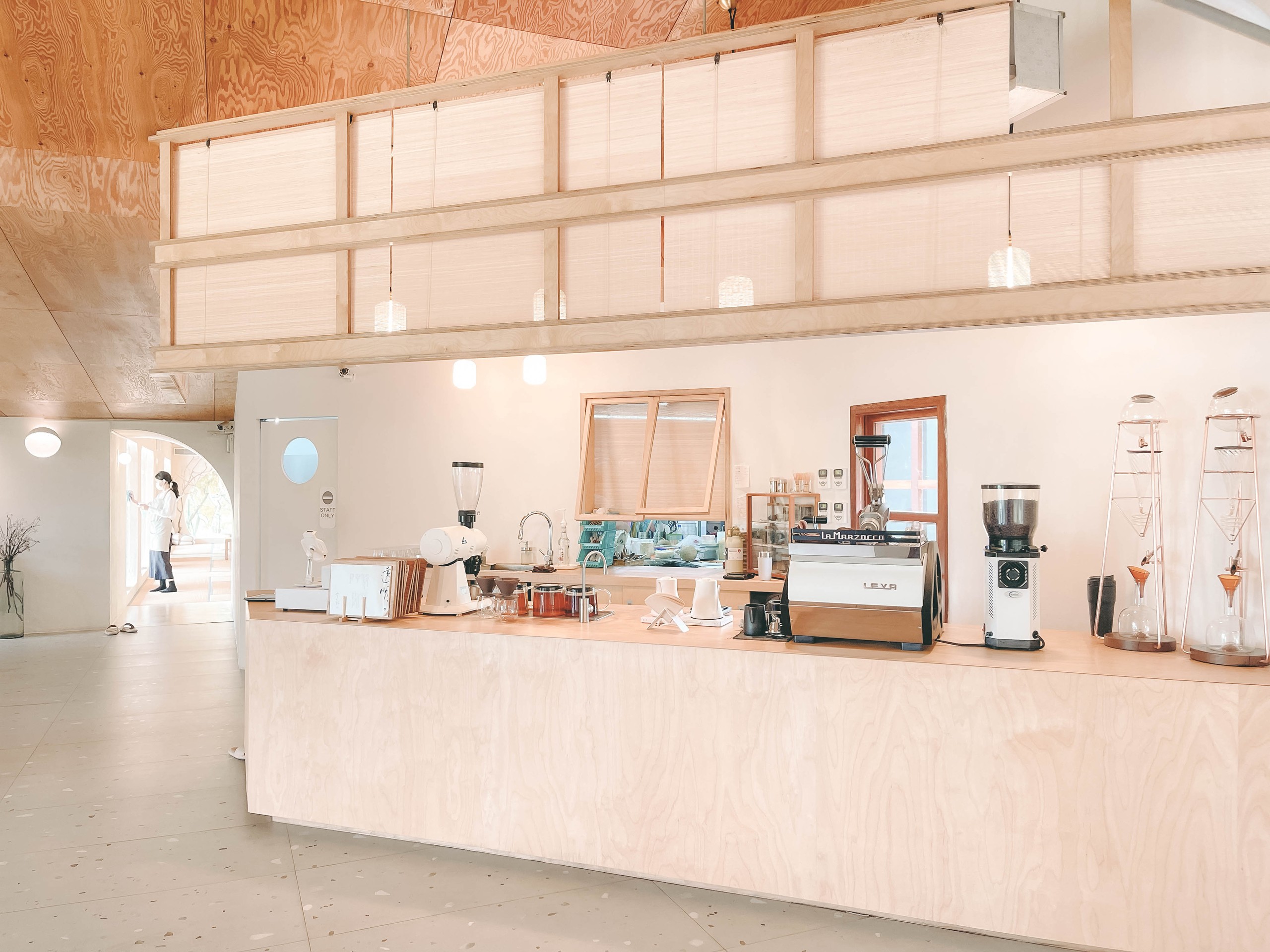 透明空氣吧台：淺色木夾板吧台與 La Marzocco 咖啡機、銅質手沖架，後方木格柵隔屏與圓窗，明亮自然光