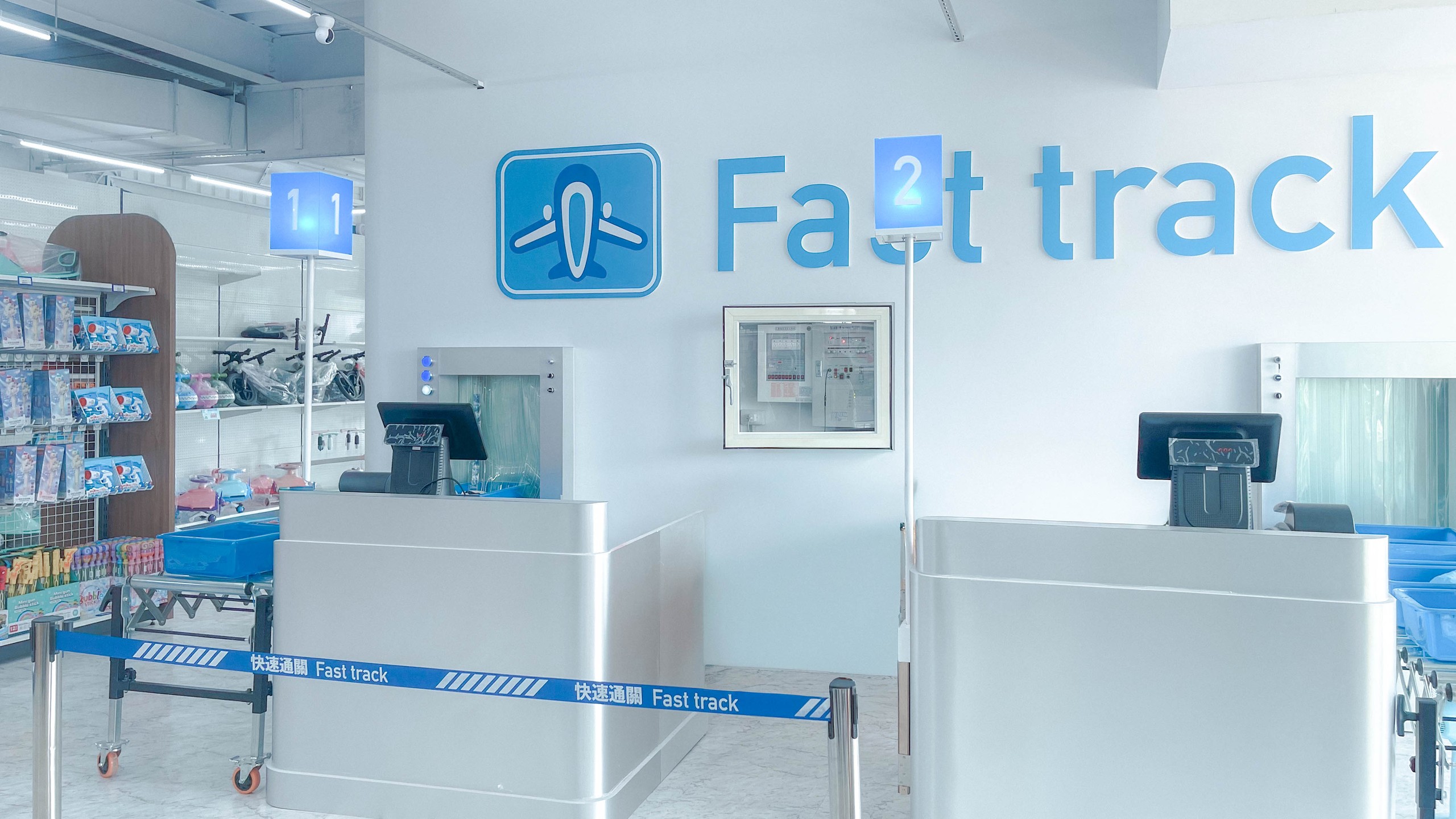 快速通關玩具店室內 Fast track 通關區：白牆上藍色飛機標誌與大字 Fast track，金屬結帳檯與藍色登機門號燈箱，像機場安檢通道
