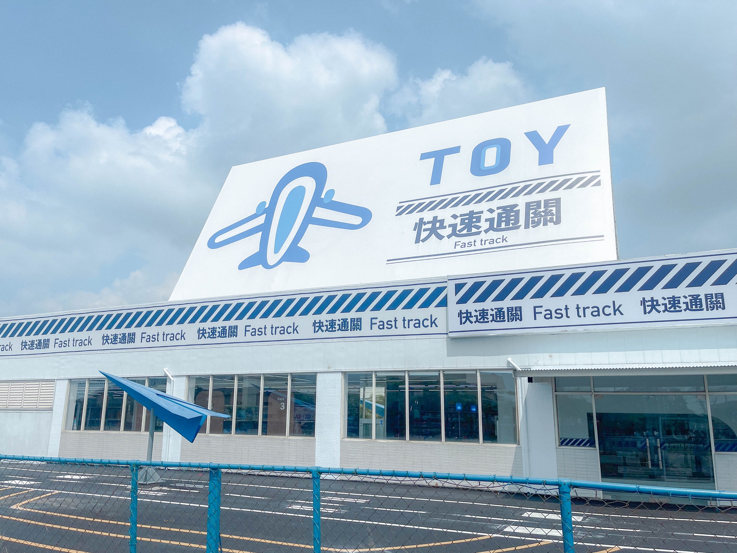 快速通關玩具店外觀，航廈造型量體與 TOY 紙飛機招牌，藍白色帶與 Fast track 字樣，像一座迷你機場